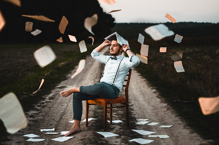A man sits on a chair in the middle of a road with book pages fluttering down around him